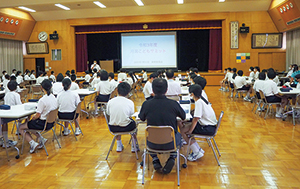 机を囲むように座り、グループを作っている中学生と大人がステージに注目している写真