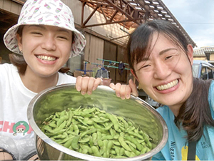 南区を訪れた人と前田さんがザルに入ったたくさんの枝豆を持ち笑顔でこちら向いている写真