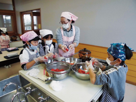 三角巾、エプロン姿の女性が三角巾、エプロン姿の子どもたちに調理実習を教えている写真