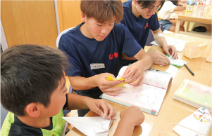 白根高校の男子生徒２人が男の子と一緒に折り紙をしている写真