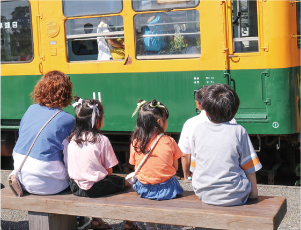 女性と女の子2人、男の子1人がかぼちゃ電車をベンチに座りながら眺めている写真