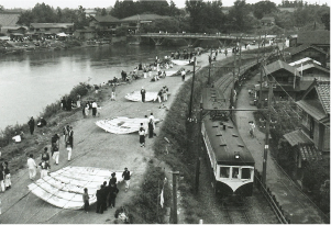 向かって左から中ノ口川・大凧が置かれた堤防・新潟交通電車線の電車と並んでいる白黒の写真