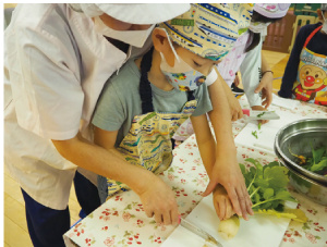 先生に手伝ってもらいながら野菜を包丁で切る諏訪木保育園の園児たちの写真