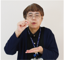 手話通訳者・鈴木さんが人差し指と親指を付けて顔の前に、反対の手は手の平を上にして胸の高さにしている写真