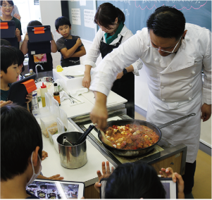 子どもたちが調理台を囲むようにし、武藤シェフが料理をしている姿を見学している写真