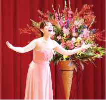 吉田早穂さんがピンクのドレスを着て、両手を広げながら歌っている写真