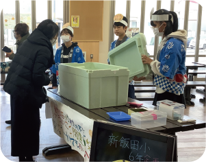 ６年生３人がお揃いの法被を着て、発泡スチロールに入っているジェラードを女性に販売している写真