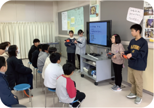 ６年生４人が教室でタブレットを使って「小林鍋」のレシピを他の生徒たちに紹介している写真
