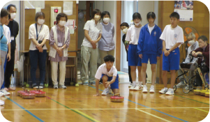 多世代交流会に参加した１年生男子がフロアカーリングの「フロッカー」を目標に向けて投げ、それを見ている人高齢者たちの写真