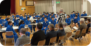 たくさんのテーブルを並べて全校生徒が車座に座り、地域の人たちの意見を聞いている写真