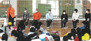大勢の小学生の前に一列に立ち、あいさつをする児童委員たちの写真