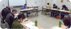 長机と椅子がロの字に配置され、委員の皆さんが着座して書類に目を通す写真