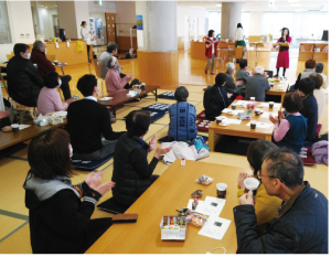 大勢の人が集まってテーブルを囲んで座り、お茶を飲みながらステージイベントを見ている写真