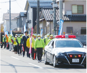 警察車両に続いて蛍光色の上着と帽子を身に着けた人たちが列になって歩く町内パレードの写真
