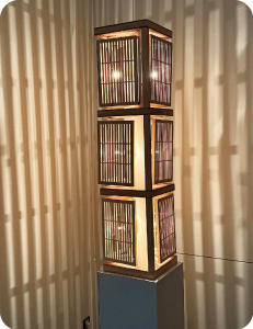 格子の板が三段に積み重なった四角柱の内側から光が照らされ、格子の影が壁に投影されている写真