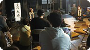 笹川邸囲炉裏の間で、囲炉裏を囲むように昔語りを聞いているお客さんの写真