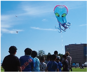 大空に揚がるタコの凧を見上げる人たちの写真