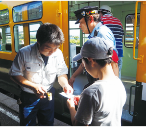 かぼちゃ電車保存会のメンバーがキャップを被ったこどもの切符を切っている写真
