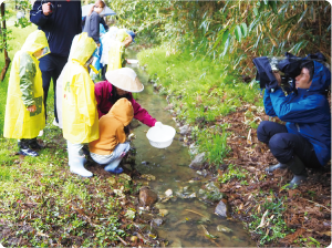雨合羽を着たこどもたちと味方ほたる愛好会が笹川邸内の小川で白い入れ物に入った「ほたるの幼虫」を放流している様子をテレビカメラが撮影している写真