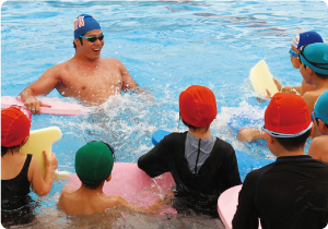 青い水泳キャップにゴーグル姿の水沼選手がプールの中でこどもたちに指導している写真