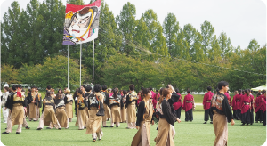 奥に大凧が揚がりその手前でよさこいを披露する団体の写真