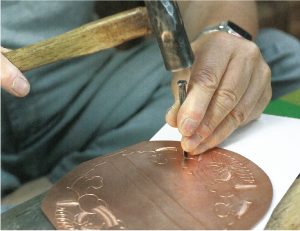 銅板に金槌を使って打ち出し作業をしている写真