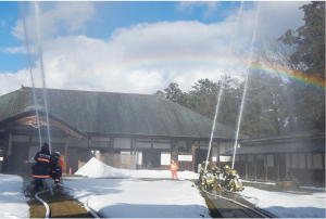笹川邸敷地内で銀色の防火衣を着た消防団員と紺色の防火衣を着た消防士が天に向けて放水訓練をしている写真