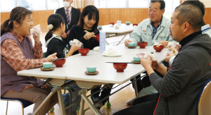 白いテーブルを囲み、女子児童と地域の男女が座り、おいしそうにおにぎりとお味噌汁を食べている写真