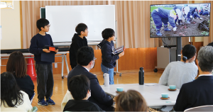 タブレットを抱えた男子児童３人が、映像が映し出されたモニターを見ながら説明している写真