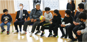 児童と地域の人たちがイスに座りながらボール送りをして楽しんでいる写真