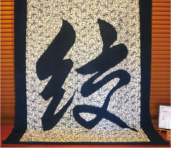 「絞」の文字が大きく描かれているタペストリー。一番外側は藍色で四角い枠があり、中央に大きく藍色の「絞」の文字があります。文字の背景は白地に藍色の細かい線が入った模様になっています