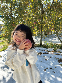 りんご畑でりんごの木をバックに両手でりんごを持ち、それを愛おしそうに目を細めながらほっぺにくっ付ける女の子の写真