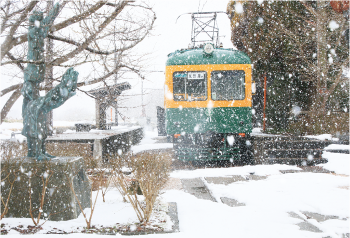 雪が降る旧月潟駅構内、左に角兵衛獅子の銅像、そして正面から見たオレンジと緑2色の「かぼちゃ電車」の写真