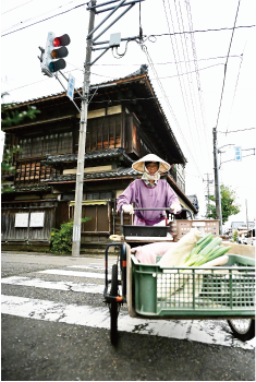 てぬぐいを被り顎で結び、その上から笠を被りピンク色の割烹着を着た女性が、白根商店街にある「庭幸酒店」前の横断歩道を野菜を乗せた押し車を押している写真