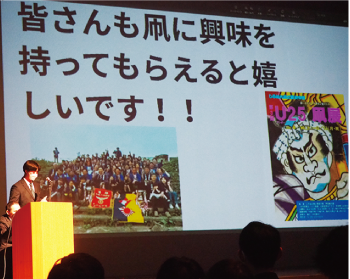 ステージ上「皆さんも凧に興味を持ってもらえると嬉しいです！」の文字とU２５凧展ポスター・昨年の凧合戦優勝組の集合写真が映し出されたスクリーンをバックに、端にある演台の前に立ち発表する男子白高生の写真
