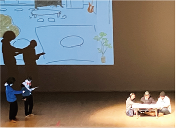 向かって左に立って演技をする白高生２人、向かって右にはテーブルを囲んで座る白高生３人が演技をするステージ上の写真