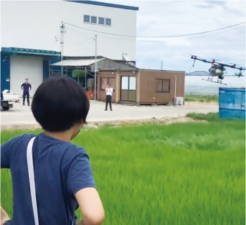 田んぼの上に飛んでいるドローンを見る味方小学校の児童の写真