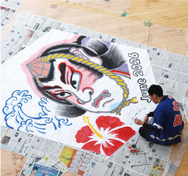 新聞紙の上に広げられた武者絵が描かれた3畳ほどの大きさの巻凧の上に、法被姿の人が胡坐をかいて座り、筆で色を塗っている写真