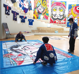 ビニールシートの上に6畳の大きさの武者絵が描かれた紙を広げ、その上に骨組みを乗せて両端を法被姿の二人が持ち、貼っている写真