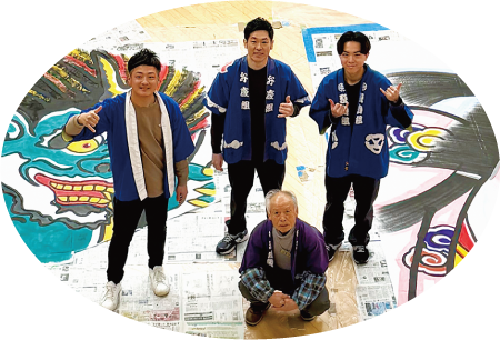 2枚の凧絵の間に座ってこちらを見ている法被姿の遠藤裕己さんと、その後列に立ってそれぞれ手でポーズをしている笑顔の富山善幸さん、尾竹一さん、山田元気さんの写真