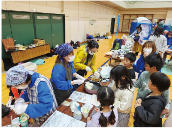 子どもたちの前でエプロン、三角巾、マスクを着けて、防災食を作っている3人の女性たちの写真