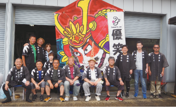 巻凧の前で黒い法被を着た人たちの集合写真