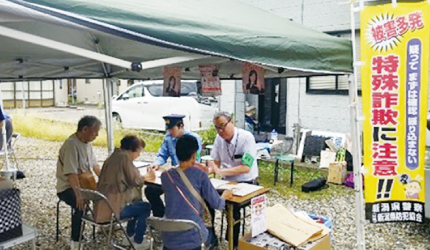 「特殊詐欺に注意?」と書かれた旗の横で警察と数名の人が座っている写真