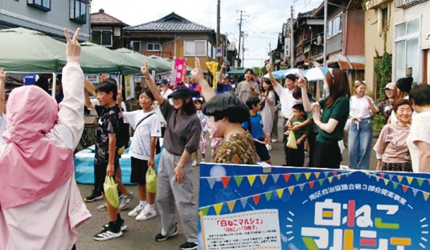 白ねこマルシェの看板の後ろでピースをして手を挙げる人たちの写真