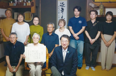 椅子に座り賞状を持つ女性と中原市長、その周りを囲む人の写真