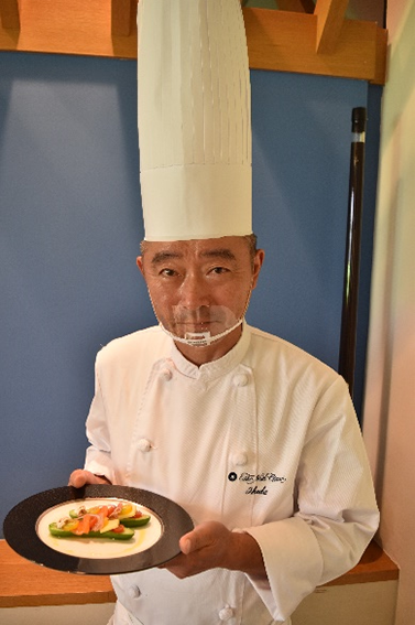 池田喜三男シェフの写真　白くて長いコック帽をかぶり手にはおいしそうな料理を持っている