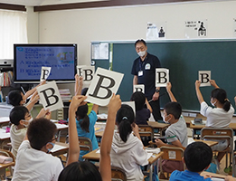 教室で講師にアルファベットが書かれたボード挙げて見せている子どもたちの写真