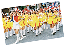黄色の衣装を着た鼓笛隊メンバーが白根本町通りを行進している写真