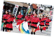 赤色の衣装を着た鼓笛隊メンバーが白根本町通りを行進している写真